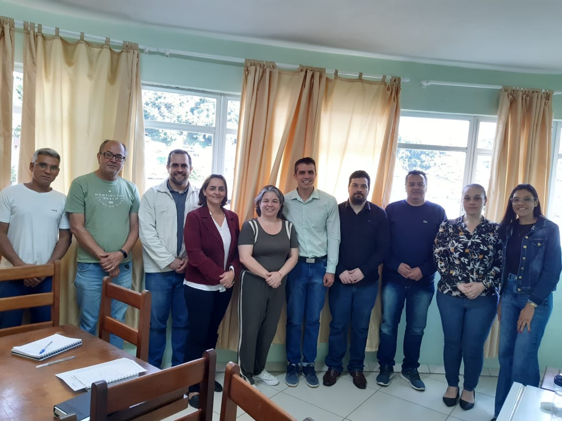 Os presentes à reunião enfileirados lado a lado, em uma sala de paredes verdes com grandes janelas. 