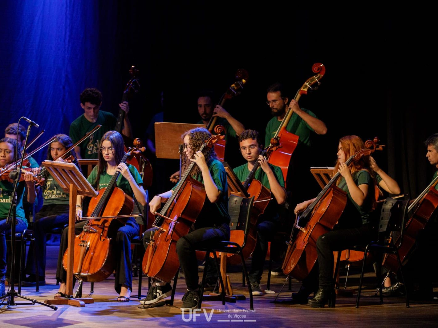 Membros da orquestra tocando violoncelos, violinos e contrabaixos num palco iluminado em azul. 