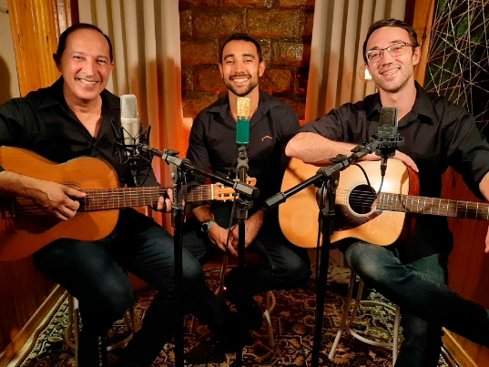 Foto do trio. Todos estão sentados, olham na nossa direção e sorriem. Dois seguram violões, um tem as mãos vazias. 