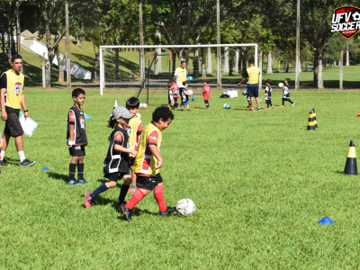 Campo de futebol em dia ensolarado, com crianças e instrutores próximos ao gol. 