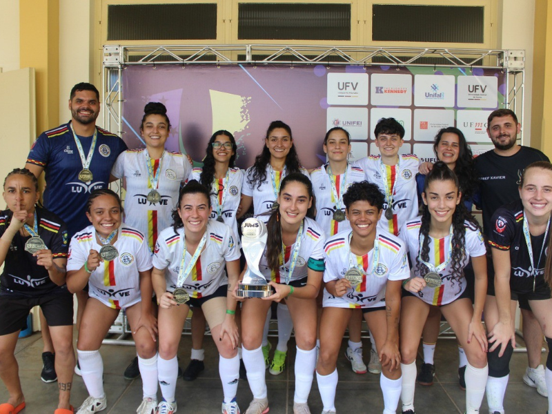 Duas fileiras de moças de uniforme branco e preto e medalhas no pescoço, e um homem em cada lado da foto. 