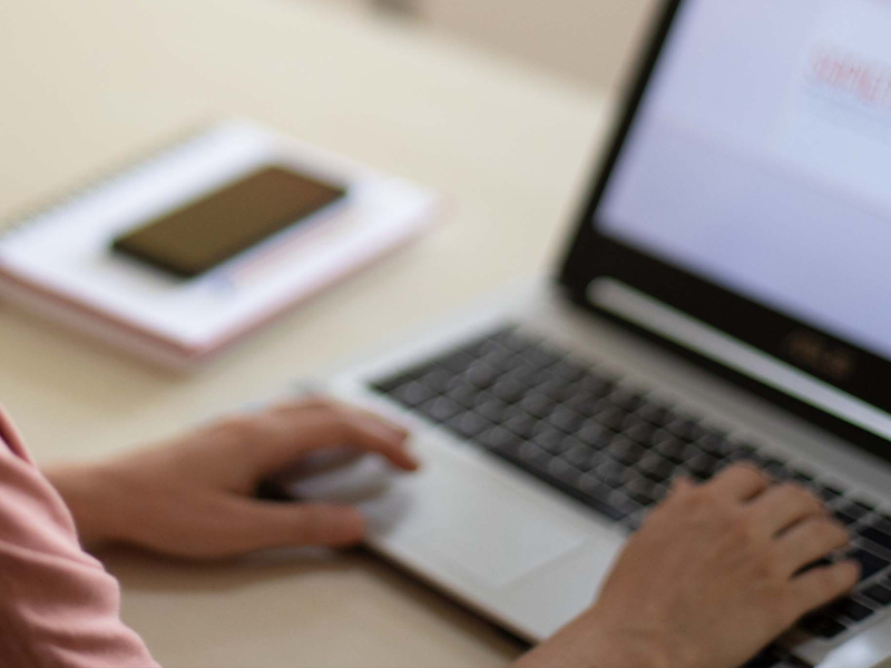Foto desfocada de mãos de pessoa branca sobre teclado de um laptop. À esquerda, caderno com um celular sobre ele. 