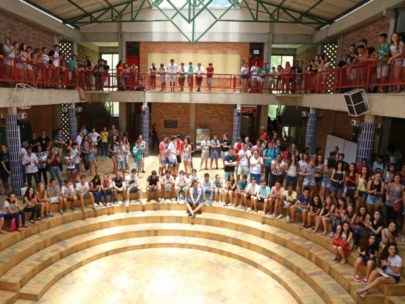 Anfiteatro bege e marrom claro com centenas de estudantes ao redor sentados e em pé, dentro de um  salão coberto. 