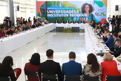 Grande mesa branca em forma de anel com todos os assentos ocupados por dezenas de pessoas. 