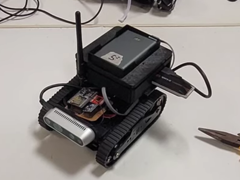 Robô de peças pretas, com uma antena em cima, uma webcam prateada na frente e duas esteias nas laterais para deslocamento. 