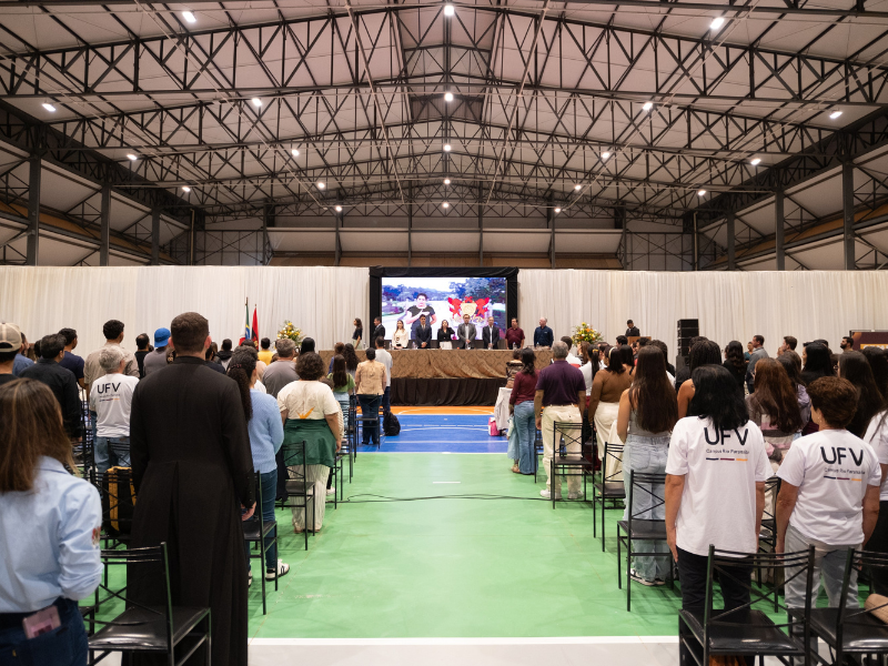 Pessoas de pé num auditório montado em ginásio. Ao fundo, mesa de abertura do evento, com telão atrás das autoridades. 