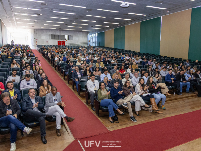 Auditório com cadeiras pretas, parcialmente ocupadas, e corredores com tapetes vermelhos. 