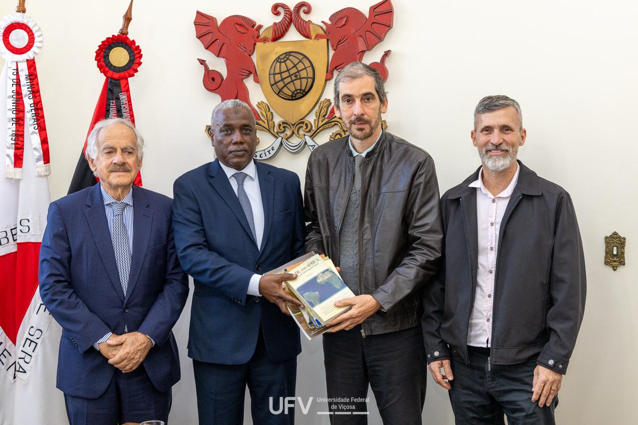 4 homens de pé lado a lado. Os dois do centro, um negro e um branco, seguram juntos alguns livros. 