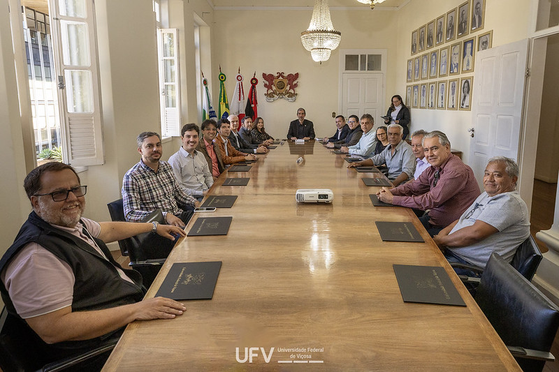 18 pessoas, entre homens e mulheres, sentados ao redor de longa mesa. Na parede ao fundo, brasão da UFV. 