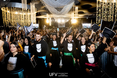 Jovens vestidos com beca de formatura e faixas coloridas na cintura vêm na nossa direção, em meio a luzes pendentes do teto. 