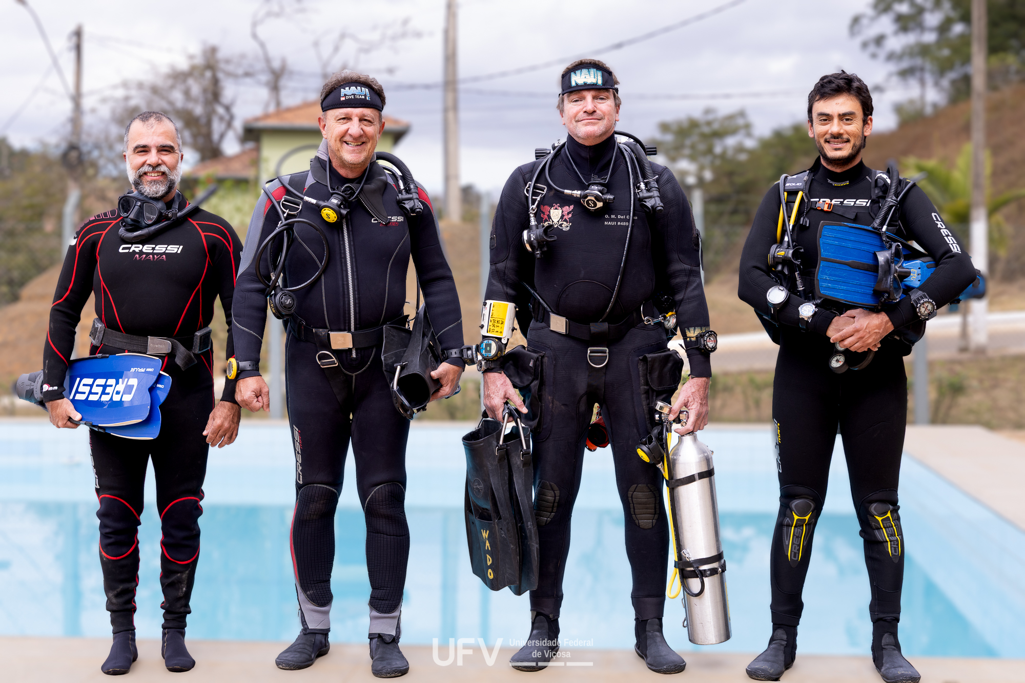4 homens de pé usando roupas e equipamento de mergulho. Estão lado a lado olhando para a frente e sorrindo. 