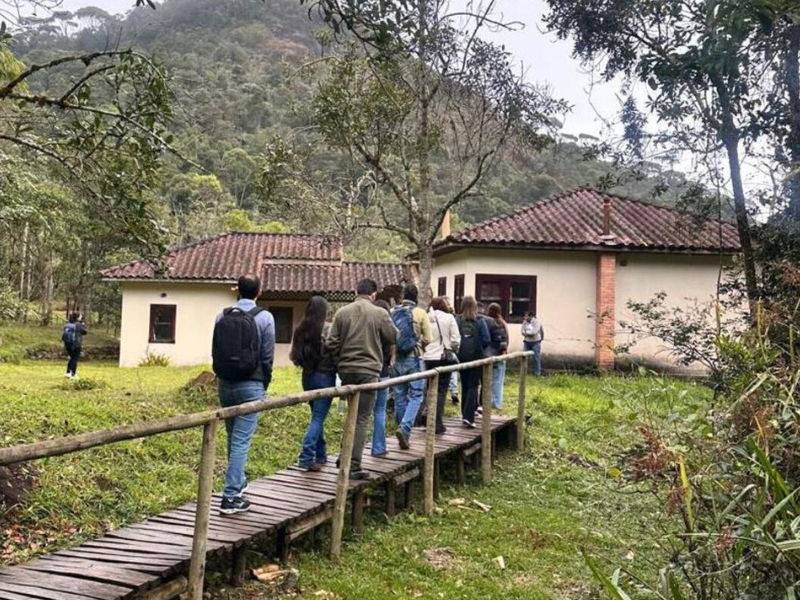 Alguns adultos jovens de calça e casaco passam por uma pequena ponte de madeira em direção a uma casinha bege. 