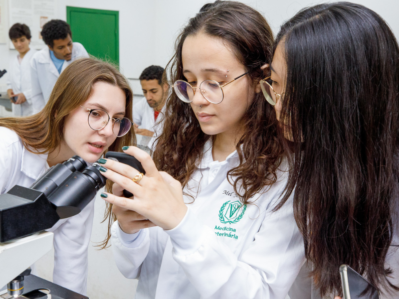 3 moças de pele clara, cabelos castanhos lisos, óculos corretivos e jaleco branco, usam celular perto de microscópio. 
