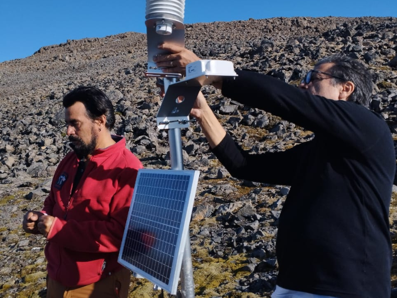 2 homens num terreno rochoso, montando um aparelho metálico com sensores e painel solar. O céu está sem nuvens. 