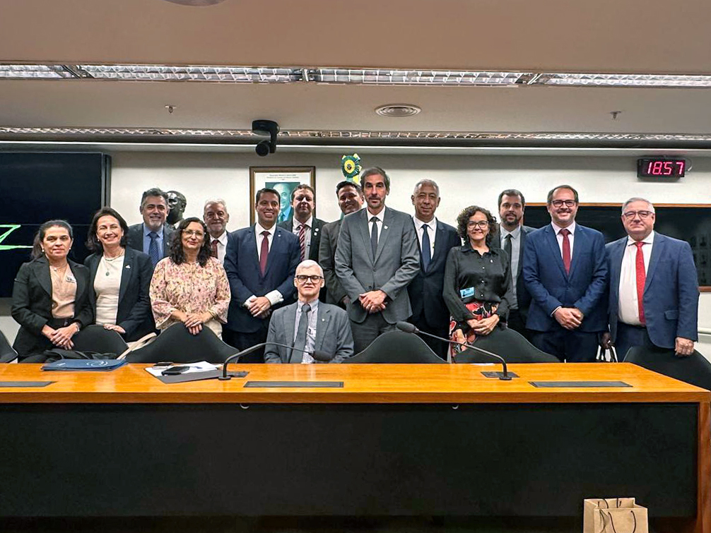 Reitor Demetrius de pé em meio a vários homens e mulheres, todos atrás de uma mesa e olhando na nossa direção. 