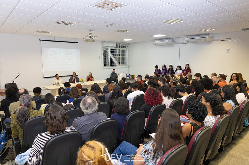 Auditório com todas as cadeiras ocupadas. Sobre o palco, há uma mesa branca com 2 homens e uma mulher. 