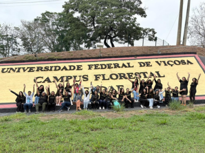 Dezenas de jovens sentados em frente a uma grande placa horizontal com 