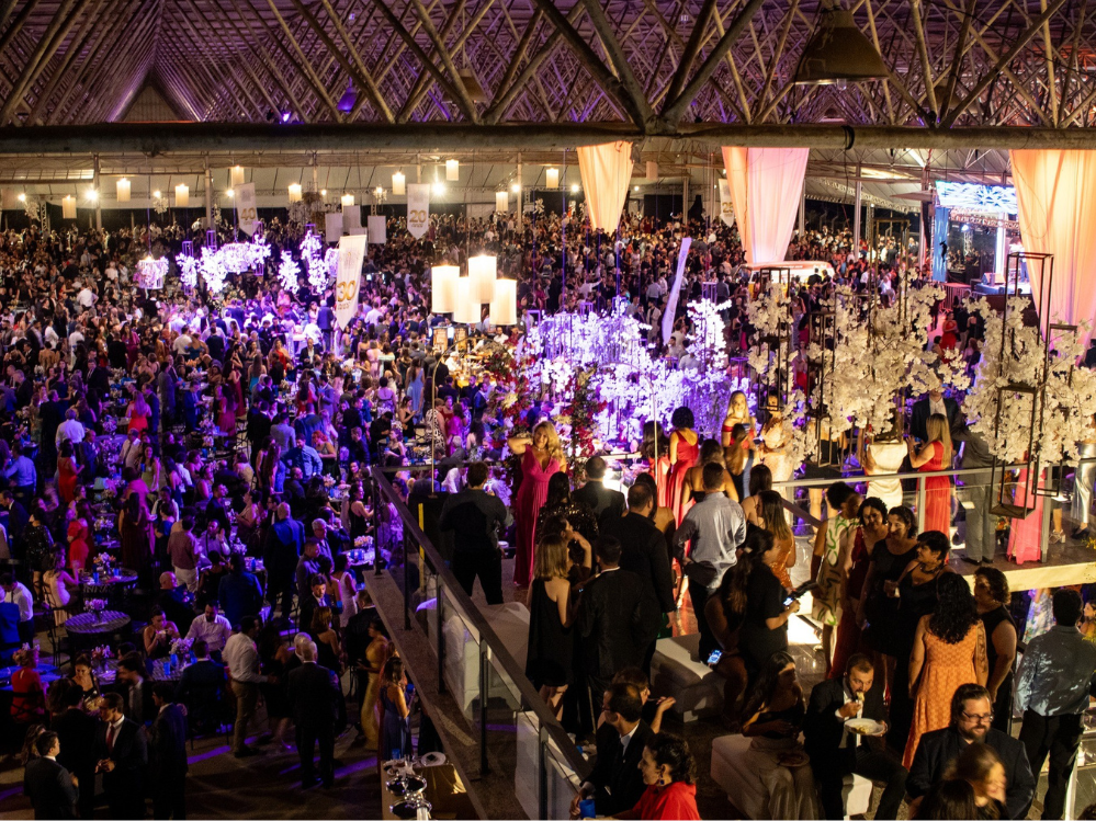 Grande salão decorado com muitos candelabros e pendentes floridos, repleto de pessoas com trajes de festa. 