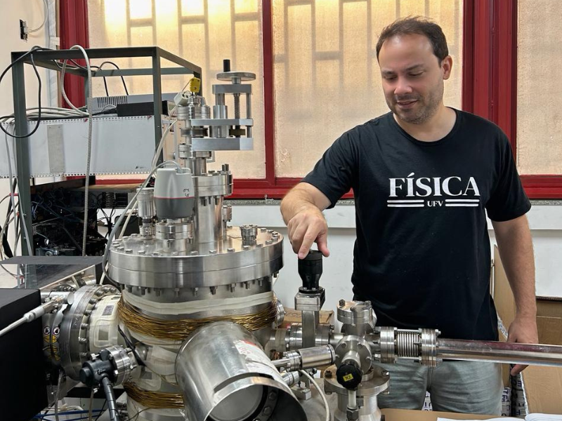 Homem ligeiramente calvo, bigode e barba por fazer, camisa da Física, mexendo em um equipamento metálico. 