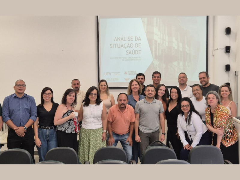 Vários homens e mulheres, de diferentes idades e etnias, lado a lado para a foto, em frente a um telão. 