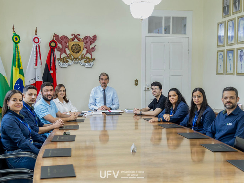 Mesa longa com 4 pessoas de cada lado, sentadas. Na ponta, homem alto, magro, de camisa branca e gravata cinza. 