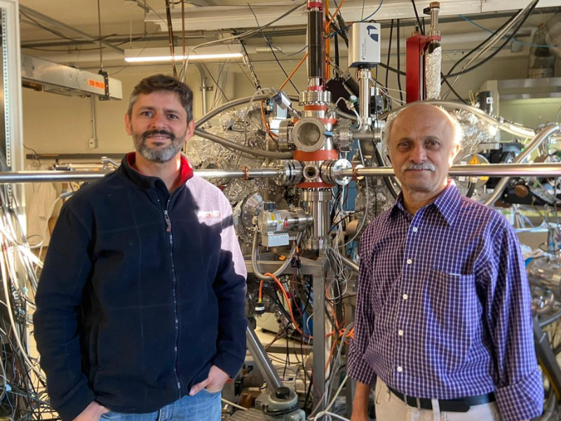 Dois homens (um de meia-idade e outro idoso) de pé em frente a um aparelho com hastes metálicas prateadas. 