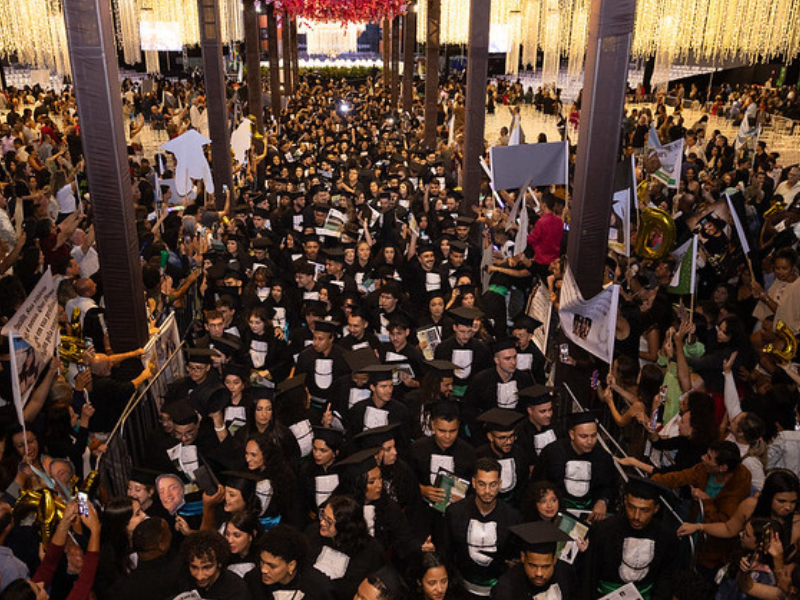 De alto a baixo da foto, centenas de formandos se caminham desordenadamente por um corredor entre colunas pretas. 