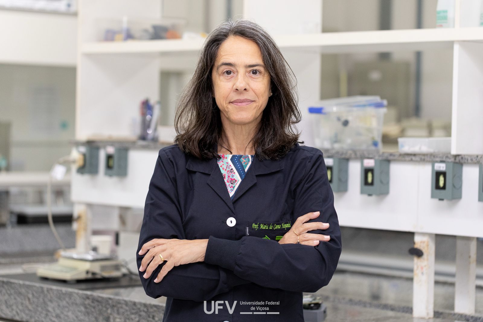 Mulher branca de meia-idade, cabelos longos escuros com raízes grisalhas, casaco azul escuro e braços cruzados. 