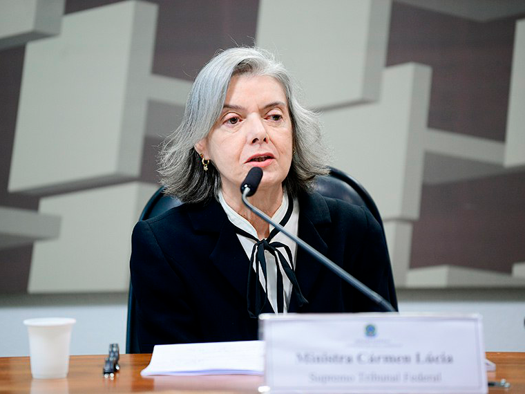 Carmem Lúcia: mulher idosa branca e magra, cabelos lisos grisalhos e olhos castanhos proeminentes. 