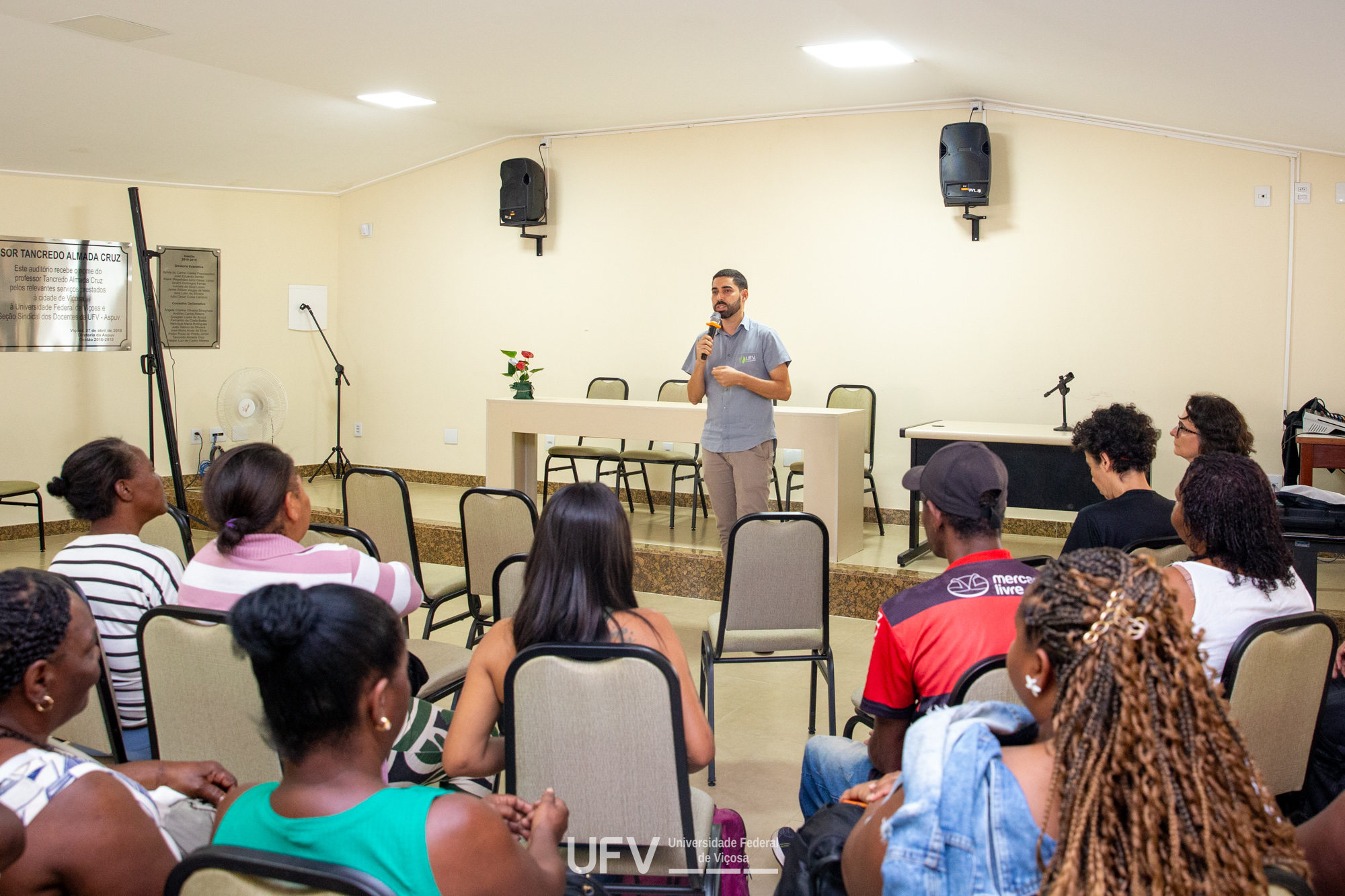Homem jovem pardo e magro fala ao microfone em um salão. Várias pessoas maioria negras, assistem sentadas. 