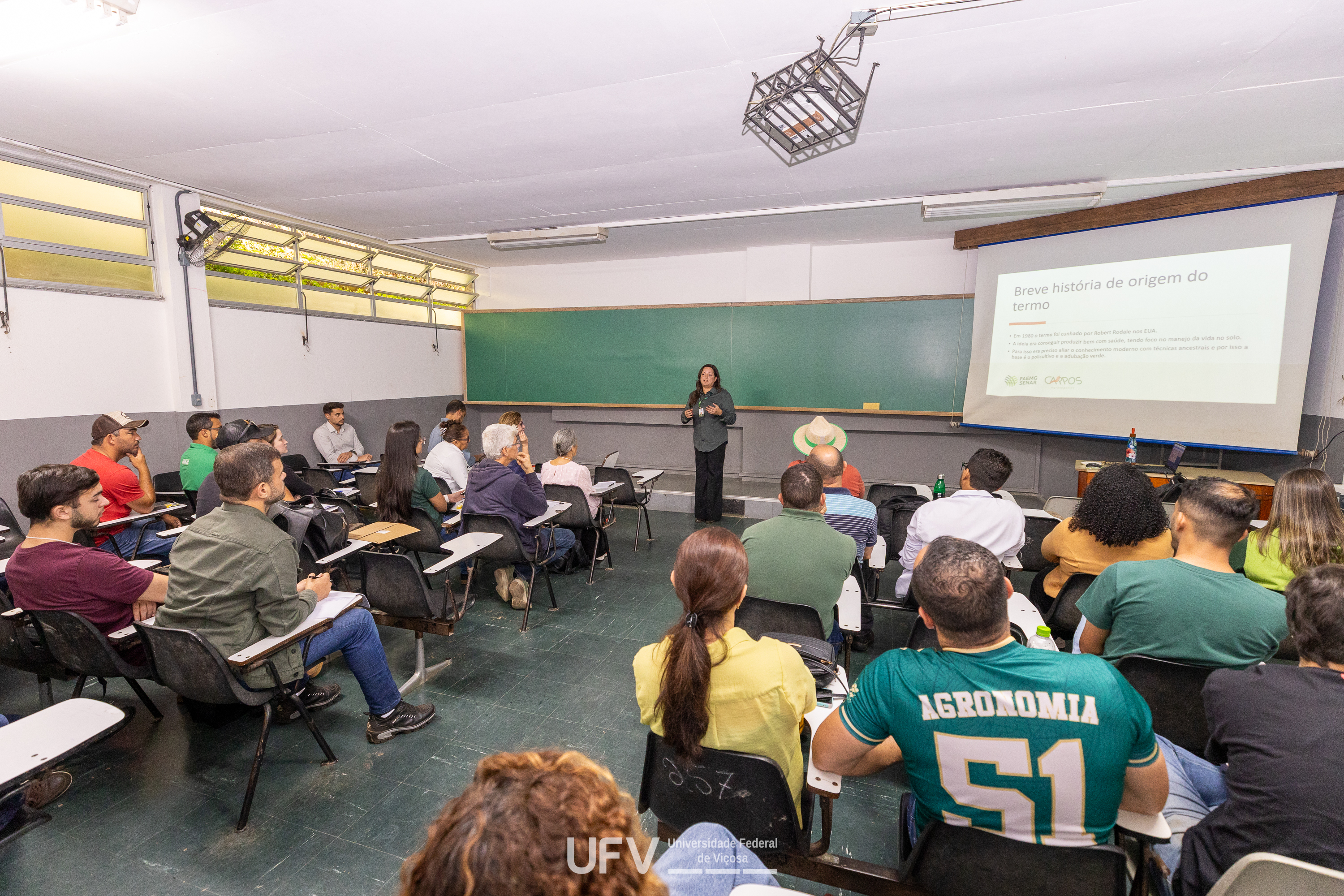 Sala de aula com alunos de diferentes idades, gêneros e etnias. Uma mulher está de pé perto do quadro. 
