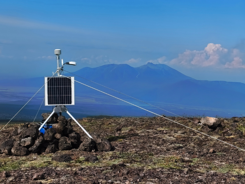 A uma grande distância, montanhas isoladas em planície. Perto, dispositivo metálico com placas solares, sobre tripé. 
