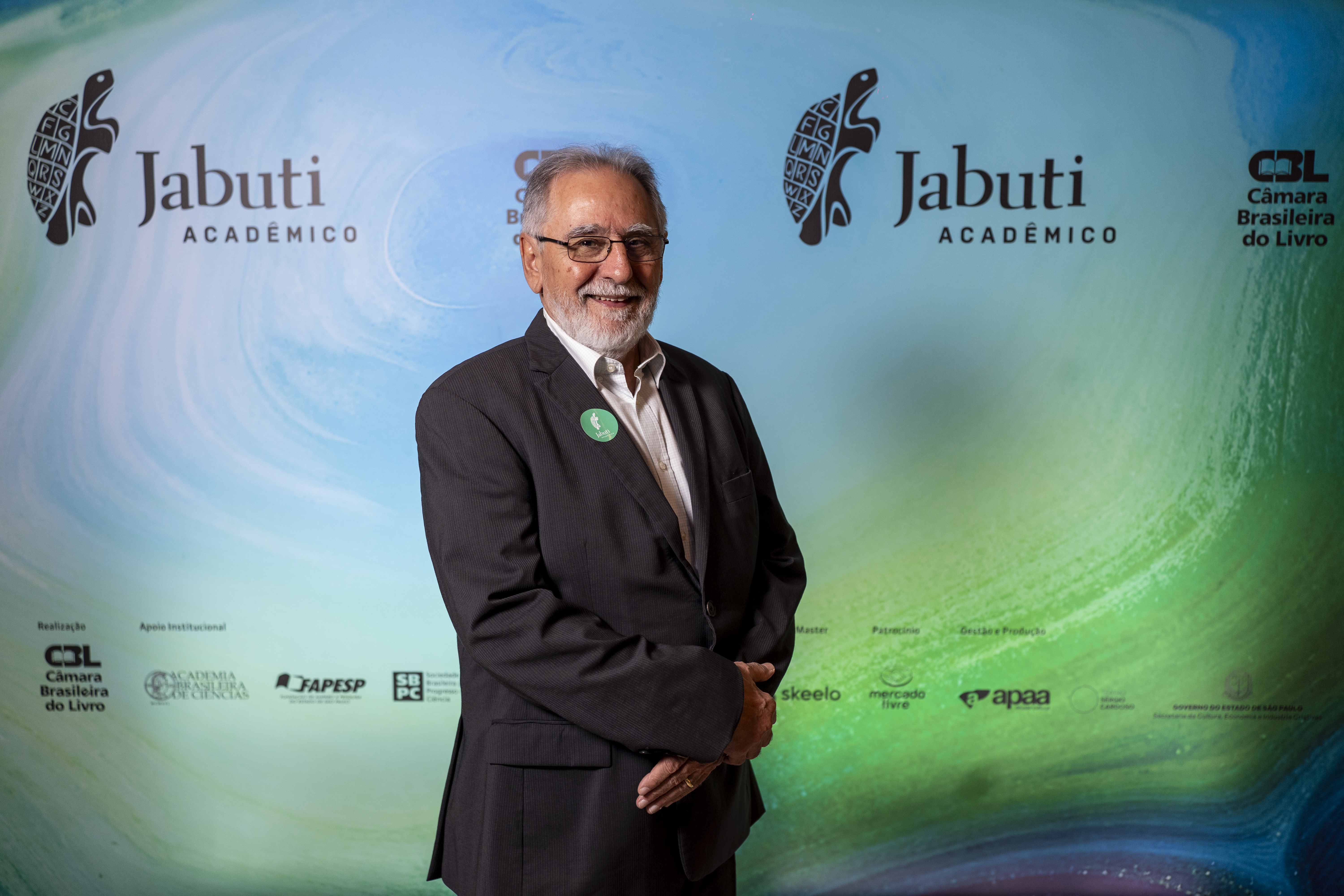 Paulo César: homem idoso, branco, cabelos grisalhos lisos, óculos e paletó preto. Olha para nós e sorri.   