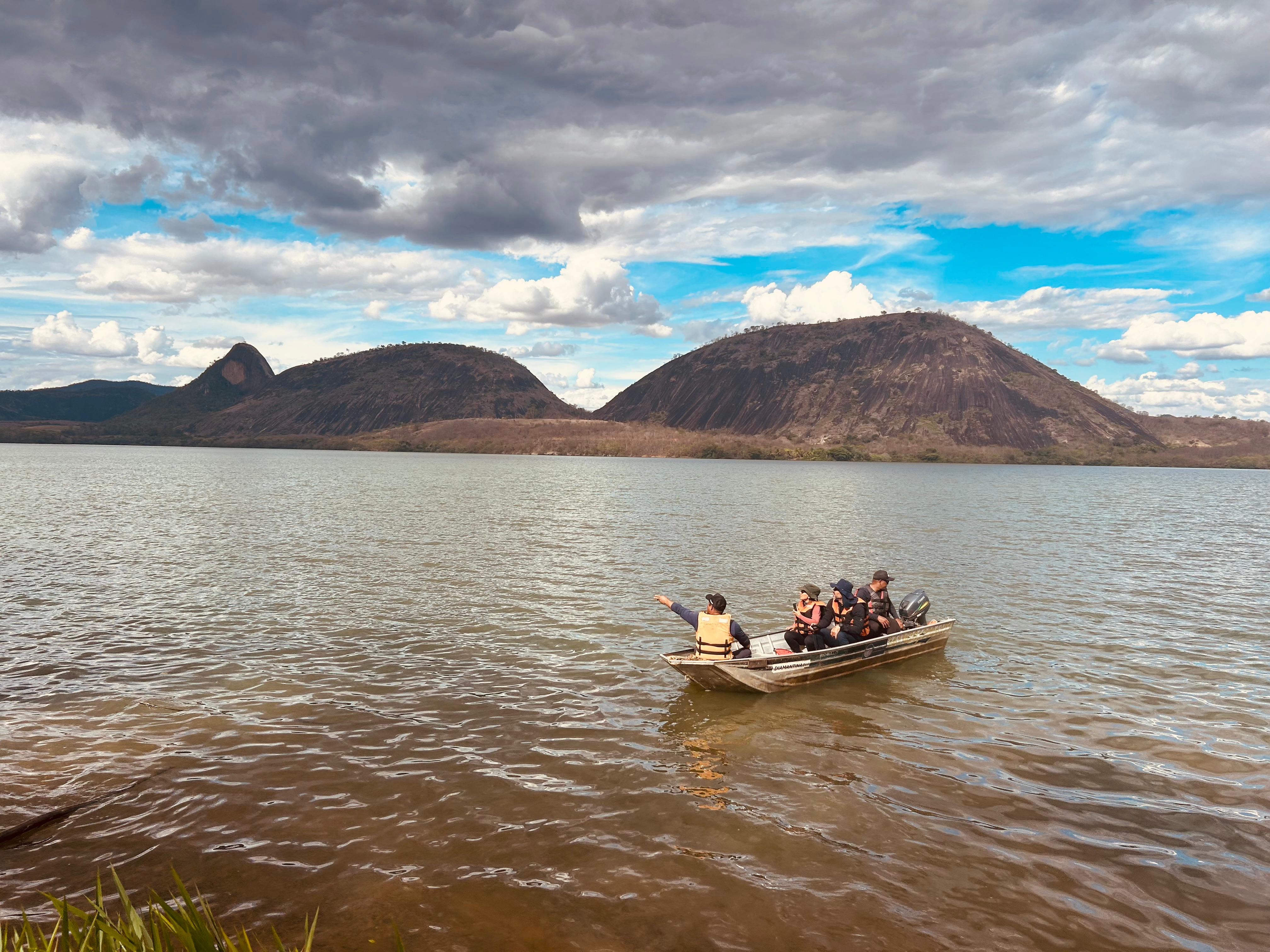 Dois homens e duas mulheres num pequeno barco em um rio largo e marrom. Ao fundo, grandes morros rochosos arredondados. 