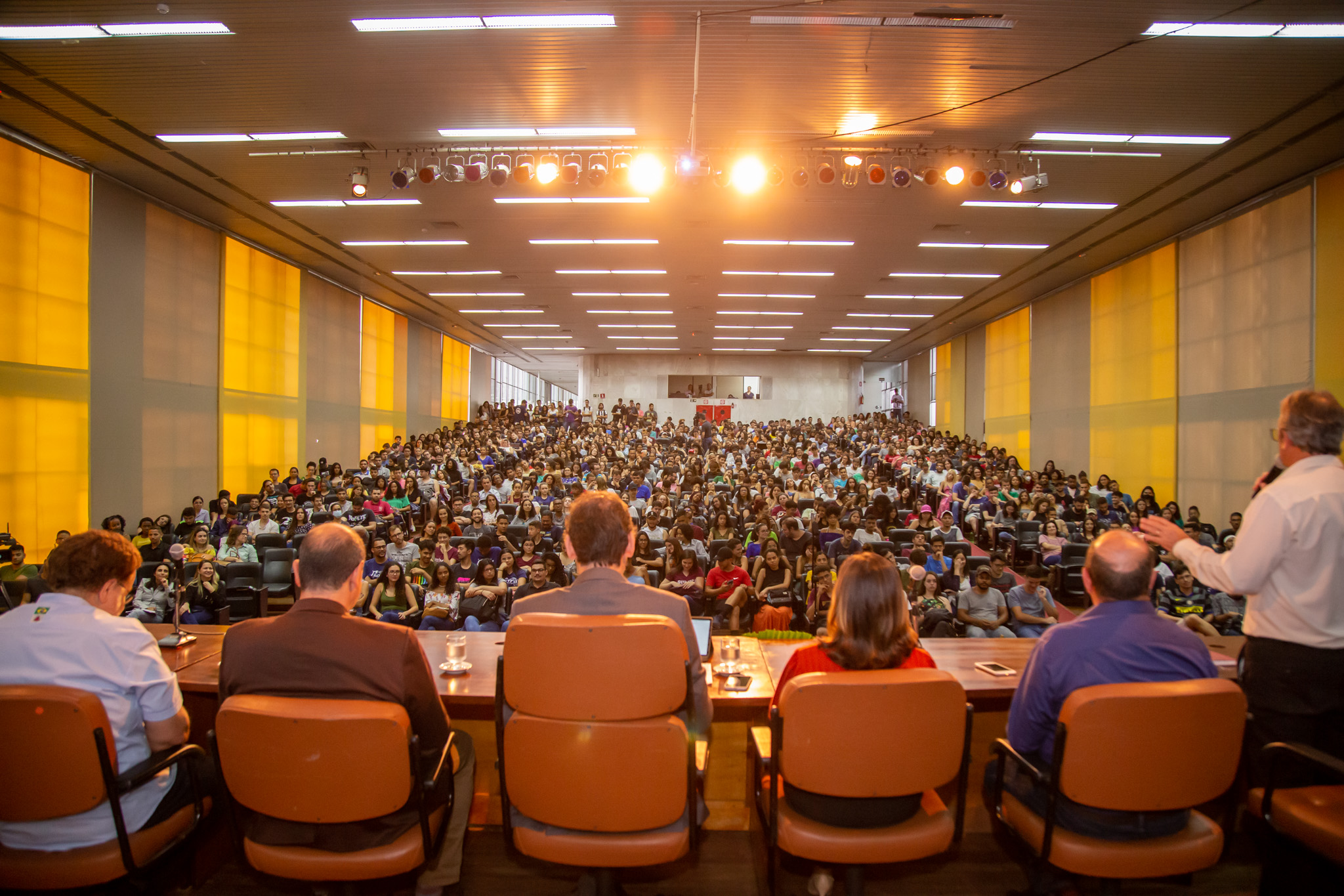 Foto tirada detrás do palco, mostrando a mesa de abertura e o auditório lotado. Alguns estudantes estão de pé ao fundo. 