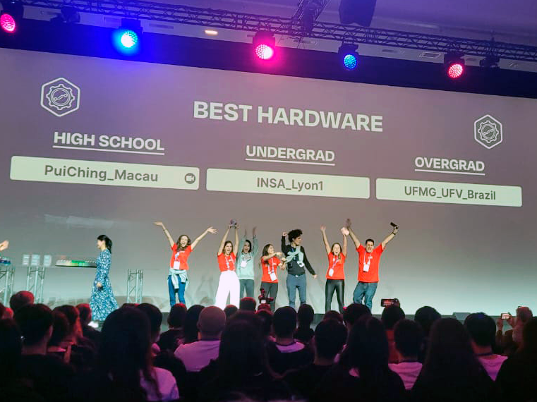 No palco do evento, estudantes da equipe vencedora comemoram após receberem o prêmio. Eles usam camisas vermelhas da equipe. 