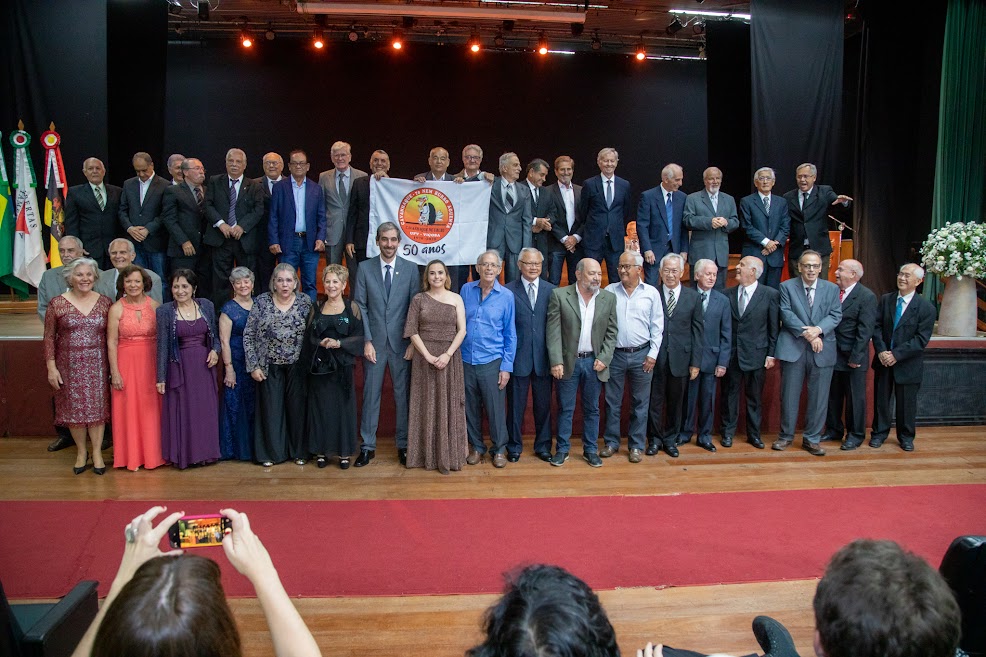 A turma de formandos “Cavanhaque de Urubu’ posa para foto no palco, com bandeira comemorativa dos 50 anos. 