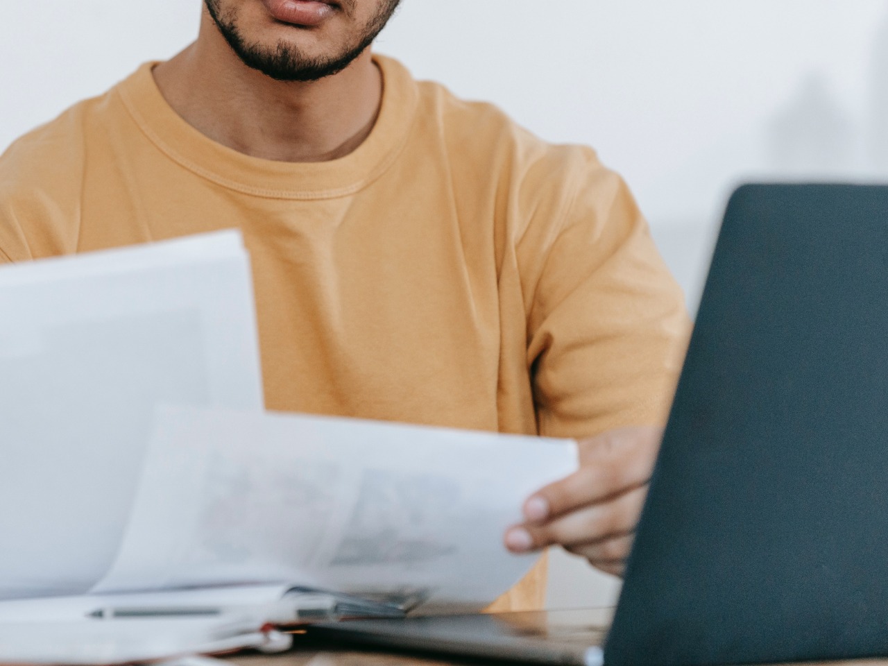 Um professor negro e com camisa laranja analisa alguns papéis. À sua frente, um laptop aberto. 