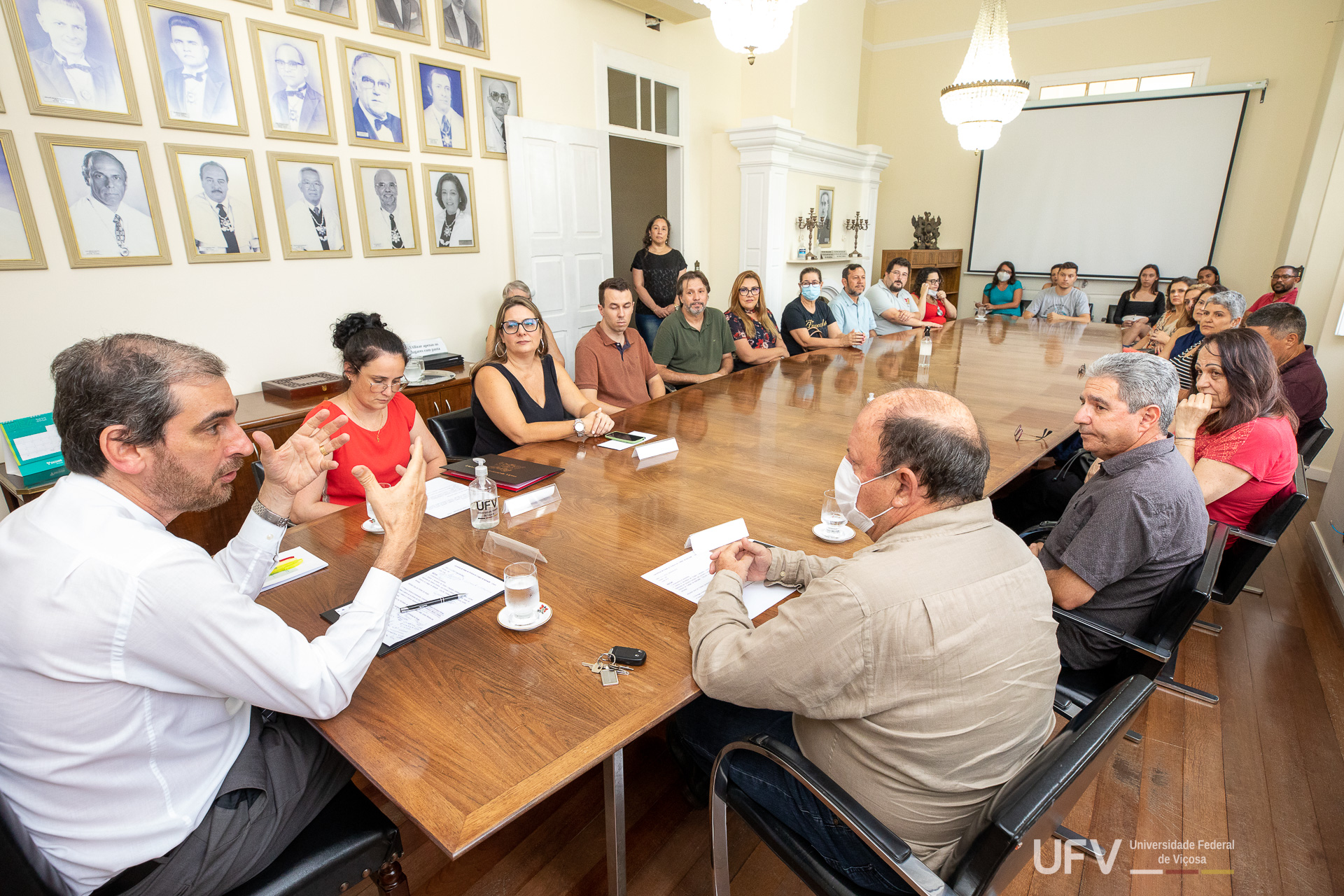 Reitor se dirige aos pró-reitores e a vários professores do Coluni sentados à mesa de reuniões. 