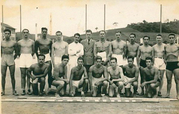 Foto antiga mostra atletas lado a lado. Ao centro, um médico e dois homens em trajes formais; os demais usam short. 