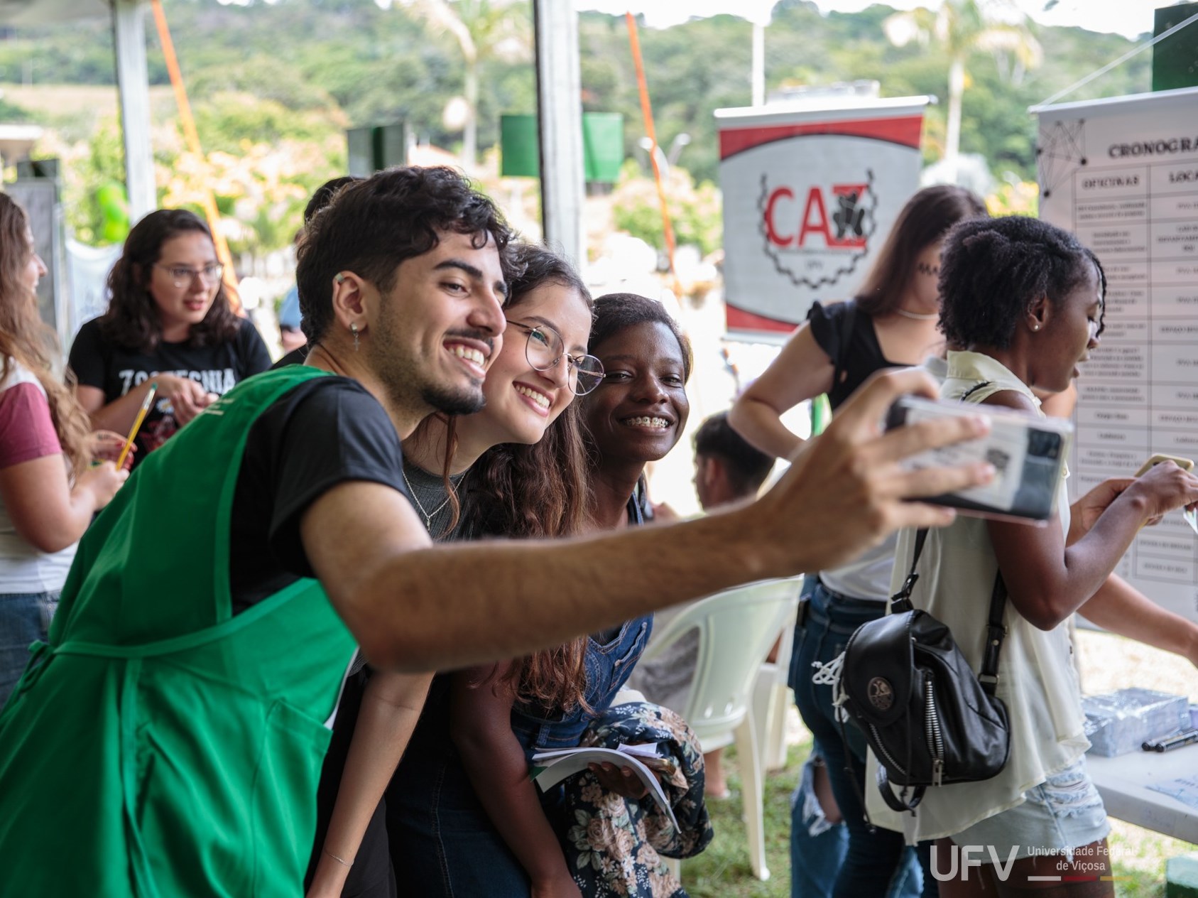 Aluno voluntário de colete verde tira selfie com calouras durante evento de recepção. 