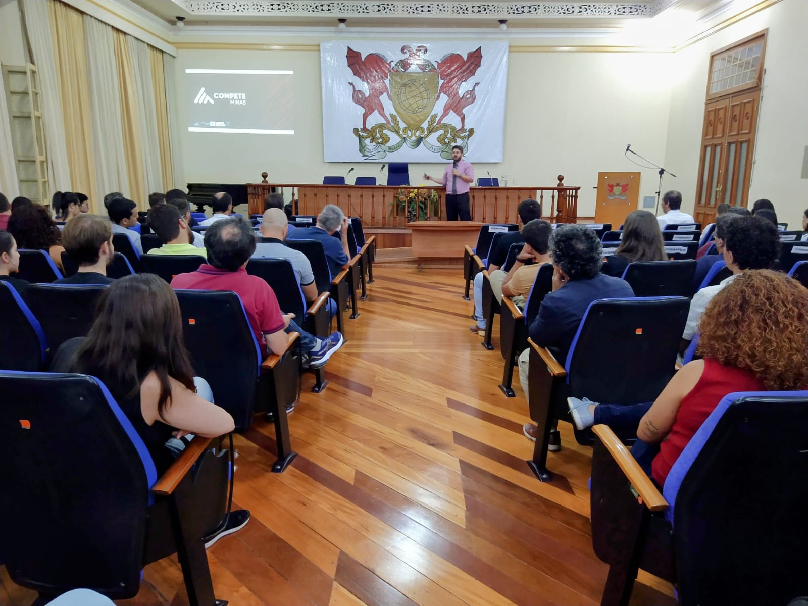 Bruno faz sua apresentação no palco do auditório com várias pessoas assistindo. Na parede ao fundo, brasão da UFV. 