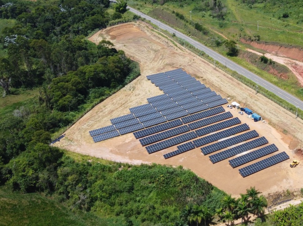 Foto aérea mostrando vários painéis solares instalados numa área verde próxima à BR120. 