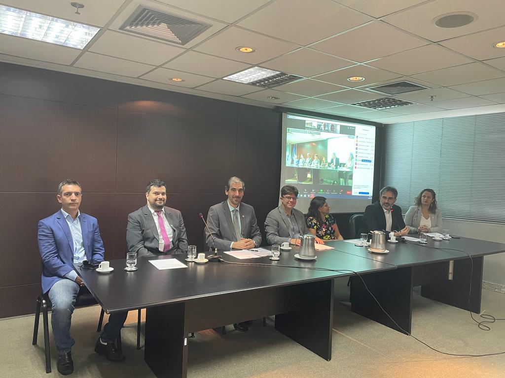 Demetrius (ao centro) e demais participantes à mesa, em frente a uma parede escura com projeção de uma tela de computador. 