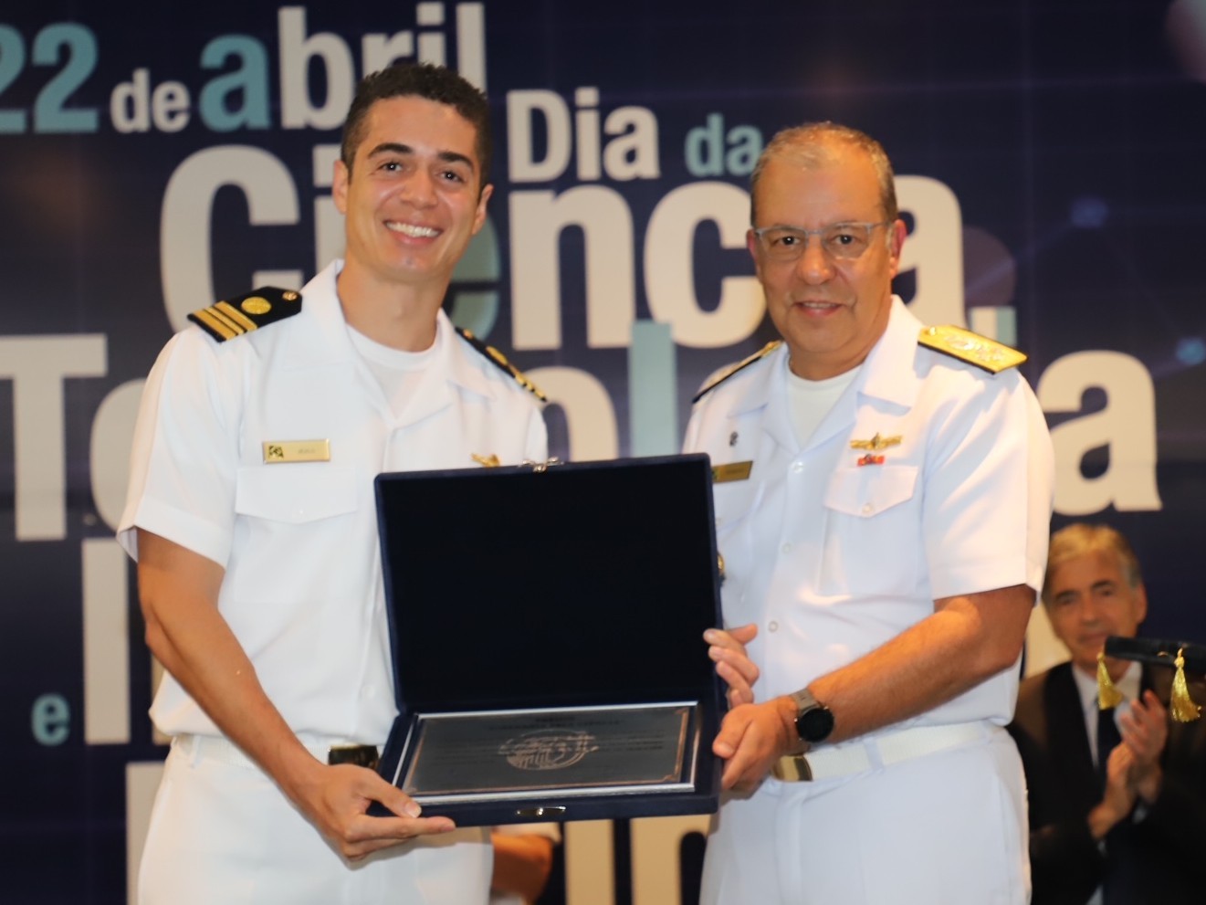 Jésus posa para a foto mostrando a placa de homenagem. Ele usa uniforme branco da Marinha. Pessoas aplaudem ao fundo. 