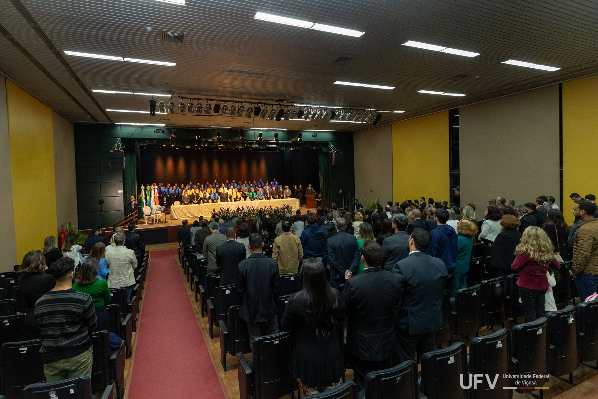 No palco do auditório, a mesa de cerimônia com todas as autoridades. Em primeiro plano, o público lota o auditório. 