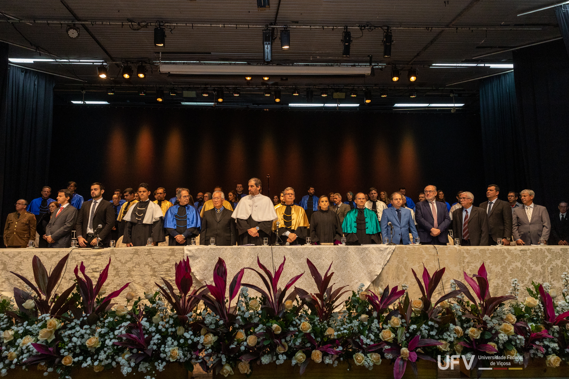 Mesa de autoridades com todos de pé durante a execução do Hino Nacional. Em frente à mesa, arranjos de flores. 