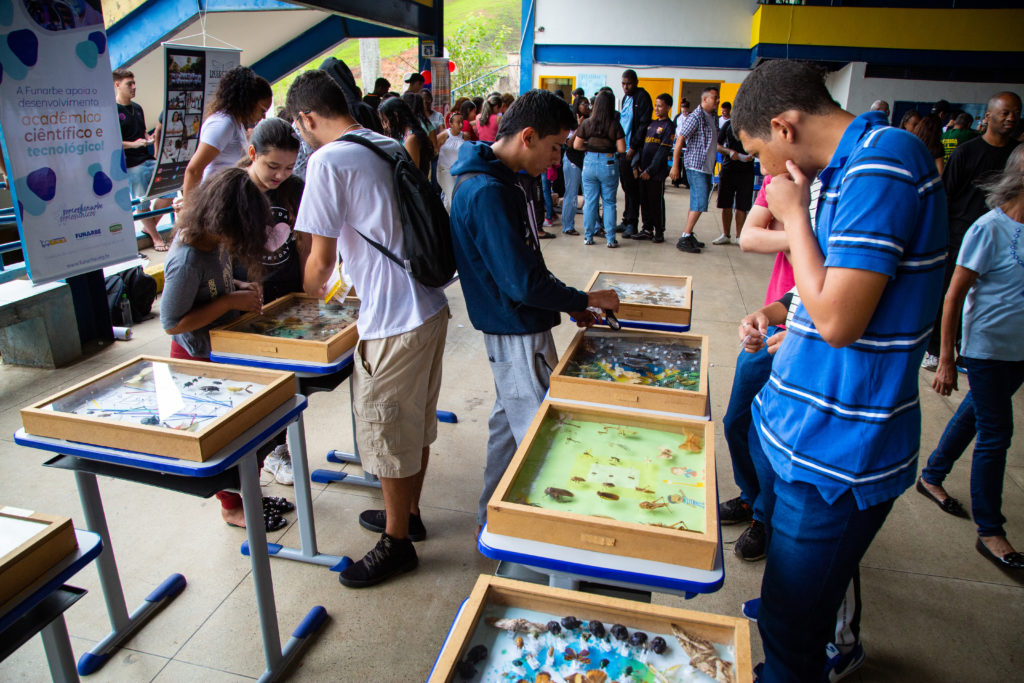 Amplo salão onde muitos alunos caminham entre trabalhos científicos. Um deles observa uma coleção de insetos. 