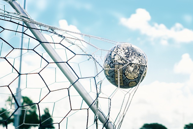 Bola de futebol no momento em que alcança a rede do gol. Ao fundo, céu azul com poucas nuvens. 
