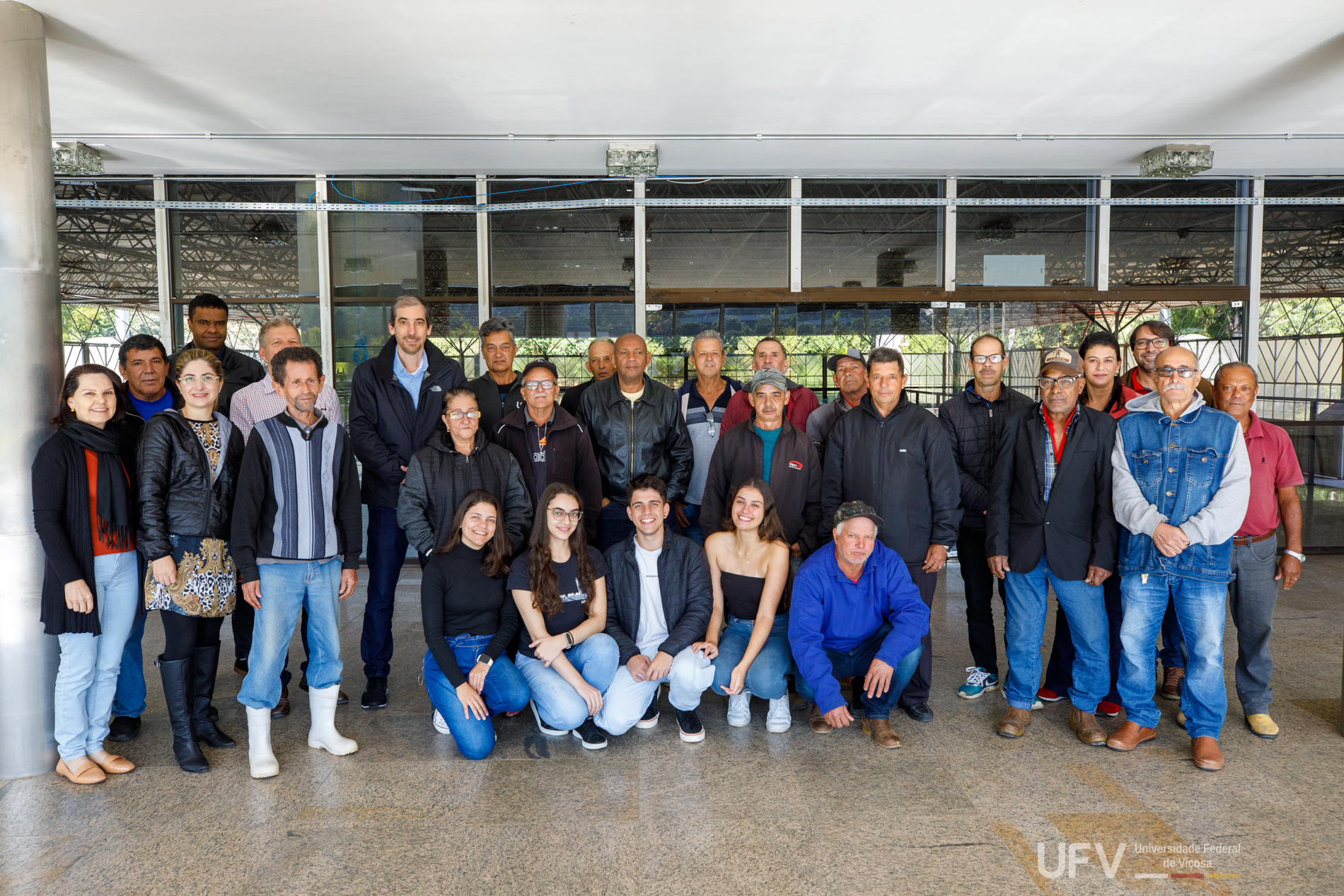 Participantes do curso alinhados lado a lado para foto, em frente a uma parede de vidro. Junto deles, o reitor da UFV. 